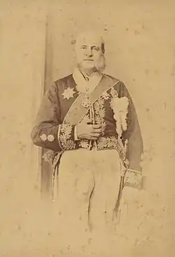Photograph of an older man with long sideburns standing in a heavily embroidered cutaway jacket adorned with various medals and wearing a sash of office