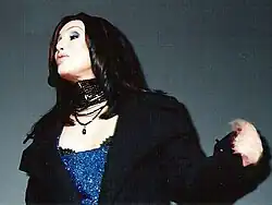 Cher performs in a glittering blue outfit with a black coat and layered necklaces, with sleek, straight dark hair.
