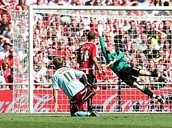 A football player in a red and white shirt and a football player in a light blue shirt are watching the ball approaching the goal, while the goalkeeper is diving to his right-hand side