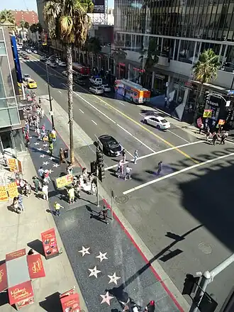 High angle looking east on Hollywood Boulevard from the north side of its intersection with Orange Drive, with palm trees, pedestrians, advertising for Madame Tussauds, and two rows of stars visible on the north side of the street. Westbound traffic is most empty, eastbound traffic is busy, and pedestrians are crossing Hollywood at its intersection with Orange. Buildings line the south portion of the street.