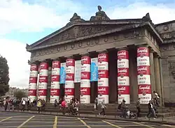 A large stone building with eight columns wrapped in red and white soup cans stacked three high behind several pedestrians. In the background is a blue sky with clouds.