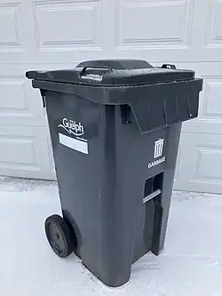A photo of a grey municipal waste bin with wheels in Guelph used to collect residential solid waste. There is snow on the ground and a white garage door in the background.