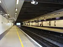 Several people walking inside a building with a black ceiling and white walls bordered in black and displaying several advertisements