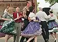 A group of dancers in colorful Western clothing promenading in a circle, with a man speaking into a microphone on a stage in the background.