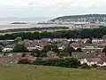 Multiple houses and other buildings around a bay into which a pier projects. On the background are hills.
