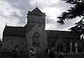 Gray stone building with arched windows. A central tower has a clock on the near face and is surmounted by a weather vane.