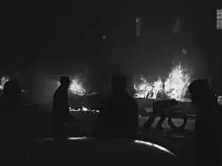 People silhouetted against cars in the background, which are engulfed in flames. One person is bent over and holding a doughnut-shaped object.
