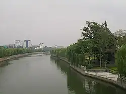 A canal scene: on the left bank stands a small pavilion surrounded by willows and other trees; along the right bank runs a row of willows, with high-rise buildings in the background