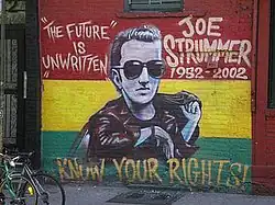 A photograph of the painting of the memorial mural of Joe Strummer on the wall of the Niagara Bar in the East Village in New York City. The mural depicts Strummer (centre) surrounded by the words "THE FUTURE IS UNWRITTEN" (on the left), "JOE STRUMMER 1952–2002" (on the right) and "KNOW YOUR RIGHTS!" (bottom) on a horizontal tricolour of red, yellow and green background