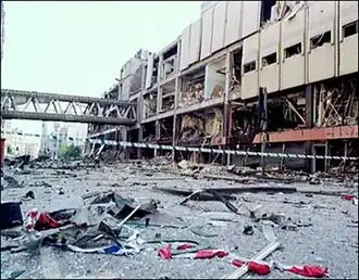 A street covered in debris. On one side is a building whose side is half-destroyed, leaving only the steel frame. The road is wet and there is crime scene tap across the centre of the photo.