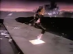 A male is shown standing in a bent down position on his toes on top of an illuminated tile. He is wearing a black jacket and pants with white shoes and a pink shirt. Behind the male a grey narrow path can be seen as well as buildings in the far background.