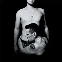 A black-and-white photo of two shirtless men. Both are wearing necklaces (one with a cross on it); one man is holding the other by the torso.