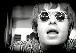 Liam Gallagher looks into the camera with lights reflecting off his glasses, in the black and white music video.