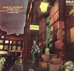 A man with blonde hair and a blue jumpsuit, holding a guitar with his foot on a higher step, dimly lit by a streetlight in an urban street
